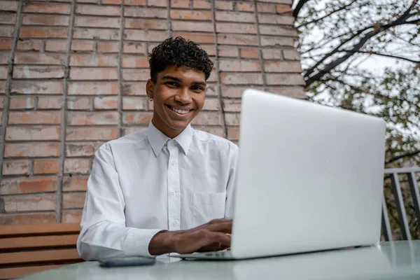 Cheerful man works at a laptop. A black guy sits on a summer terrace or veranda near a brick wall. View of the spring business downtown