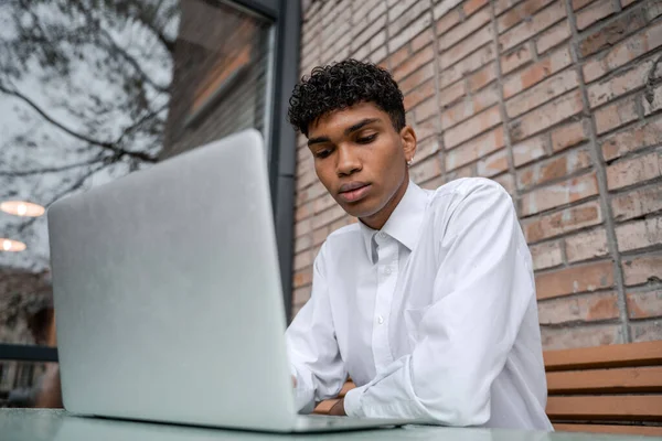Veselý muž pracuje v laptopu. Černoch sedí na letní terase nebo verandě u cihlové zdi. Pohled na jarní podnikání v centru — Stock fotografie
