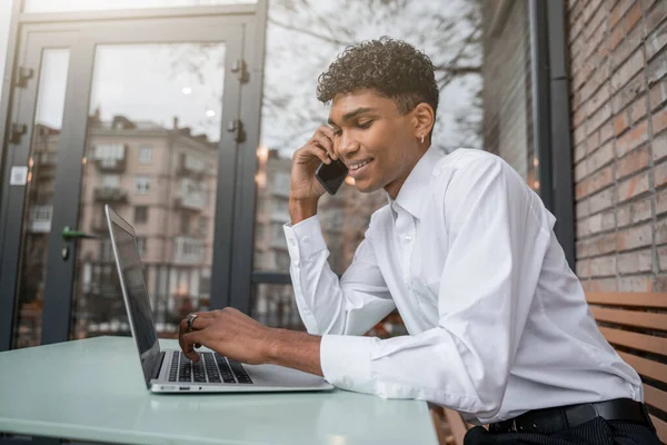 Veselý muž pracuje v laptopu a mluví po telefonu. Černoch sedí na letní terase nebo verandě u cihlové zdi. Pohled na jarní podnikání v centru — Stock fotografie