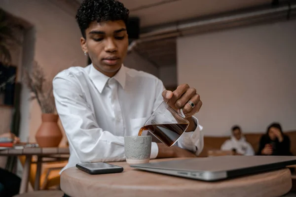 Mladý černoch nalévá čaj z konvice do šálku. Ten chlap v kavárně. Práce mimo kancelář, vzdálenou práci nebo studijní koncept. — Stock fotografie
