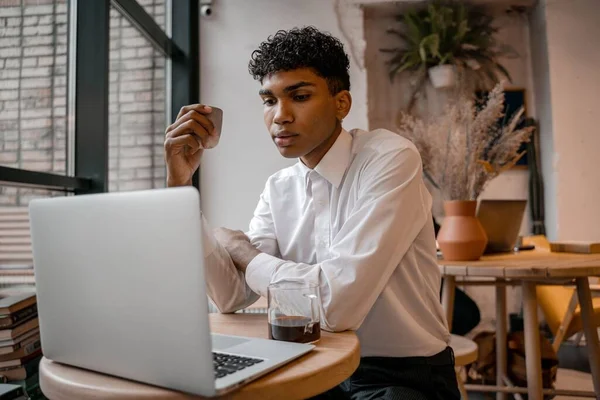 A young black man is sitting at the table with a laptop, drinking tea and using a smartphone. The guy in the cafe. Work outside the office, remote work or study concept.