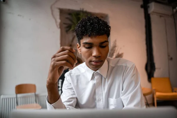 A young black man is sitting at the table with a laptop, drinking tea and using a smartphone. The guy in the cafe. Work outside the office, remote work or study concept.