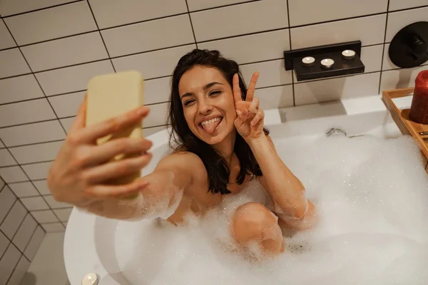 Junge Frau macht Selfie, während sie es sich in der schönen Badewanne voller Schaumstoff mit Kerzen verziert gemütlich macht. Gesunde Behandlung und Genuss — Stockfoto