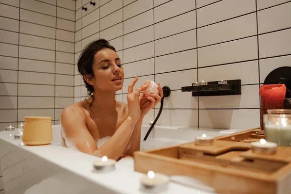 Junge Frau mit Maske auf dem Gesicht, entspannend in dem gemütlichen Bad voller Schaum und dekoriert mit Kerzen. Gesunde Behandlung und Genuss. — Stockfoto