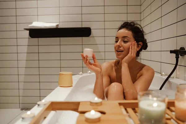 Junge Frau mit Creme im Gesicht, entspannend im gemütlichen Bad voller Schaum und dekoriert mit Kerzen. Gesunde Behandlung und Genuss — Stockfoto