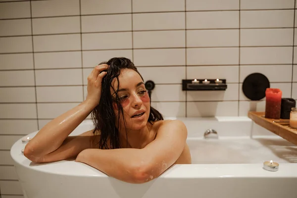 Junge und sexy Frau mit Augen- und Lippenflecken, die es sich in der gemütlichen Badewanne voller Schaumstoff mit Kerzen gemütlich macht. Gesunde Behandlung und Genuss. — Stockfoto