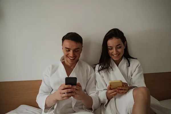 Mañana de feliz pareja joven en batas blancas en la cama en casa, usando teléfonos — Foto de Stock
