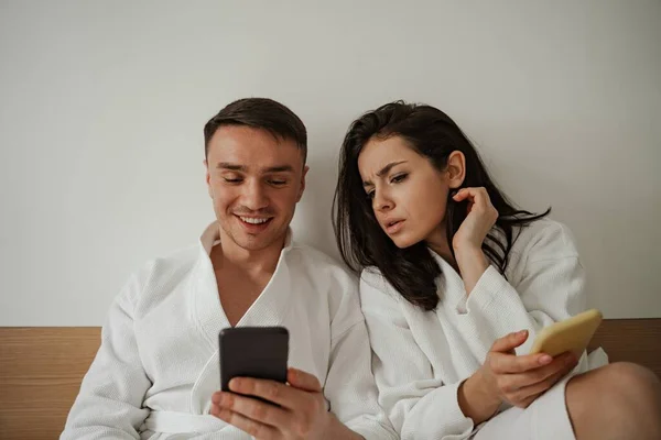 家の中の白いバスローブで幸せな若いカップルの朝、携帯電話を使用して — ストック写真