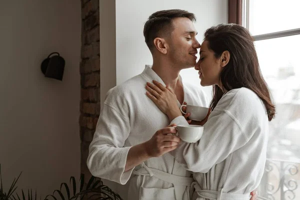 Manhã de feliz jovem casal beijando em roupões de banho brancos em casa, com xícara de café — Fotografia de Stock