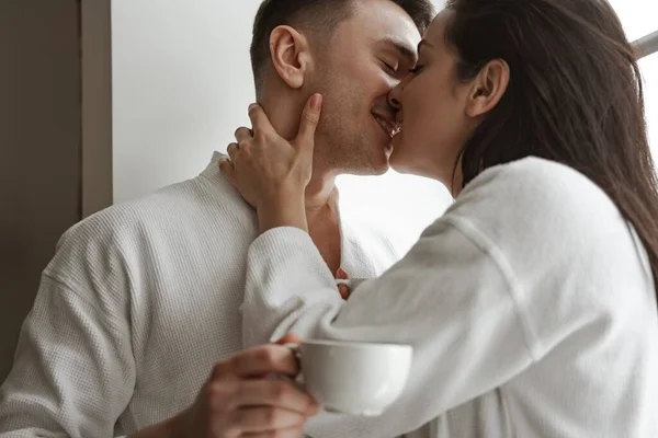 Πρωινό ευτυχισμένου ζευγαριού που φιλιέται με λευκά μπουρνούζια στο σπίτι, με ένα φλιτζάνι καφέ — Φωτογραφία Αρχείου