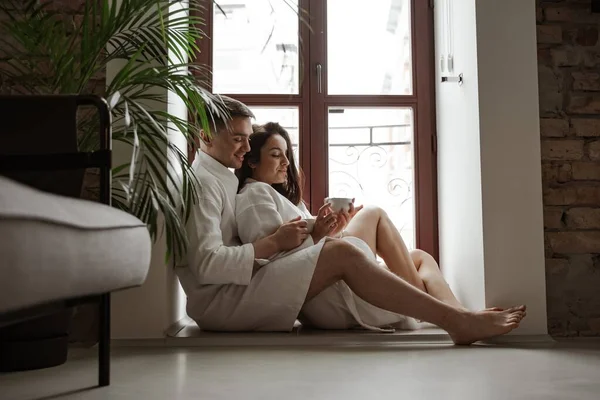 Ранок щасливої молодої пари в білих халатах вдома сидить на вікні, п'є каву і обіймає — стокове фото