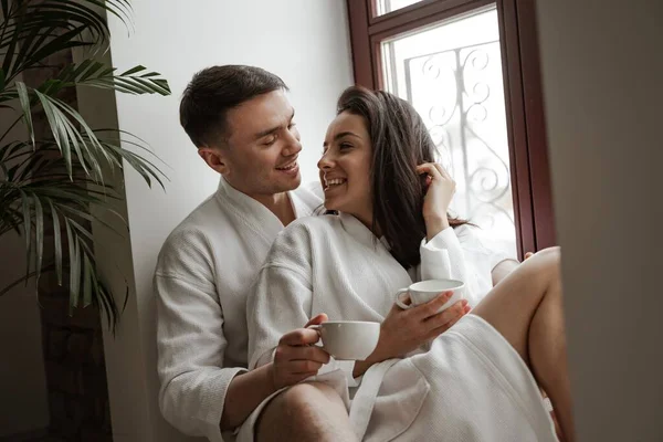 Manhã de jovem casal feliz em roupões de banho brancos em casa sentado na janela, bebendo café e abraço — Fotografia de Stock