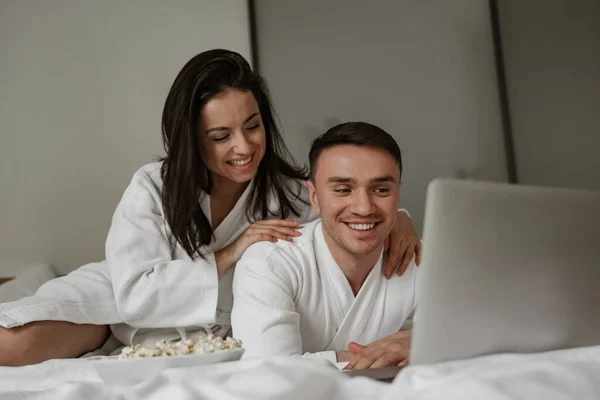 Feliz pareja joven en batas blancas en casa en la cama con maíz pop, viendo películas en el ordenador portátil y riendo — Foto de Stock