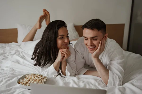 Ευτυχισμένο νεαρό ζευγάρι με λευκά μπουρνούζια στο σπίτι στο κρεβάτι με ποπ κορν, βλέποντας ταινία στο laptop και γελώντας — Φωτογραφία Αρχείου