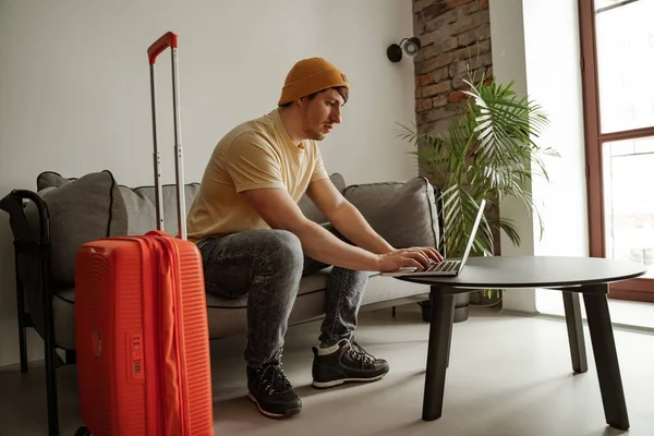 Hombre joven viajero está sentado en el sofá utilizando el ordenador portátil en la habitación de hotel en las vacaciones de verano. Hombre fresco en ropa casual con equipaje — Foto de Stock