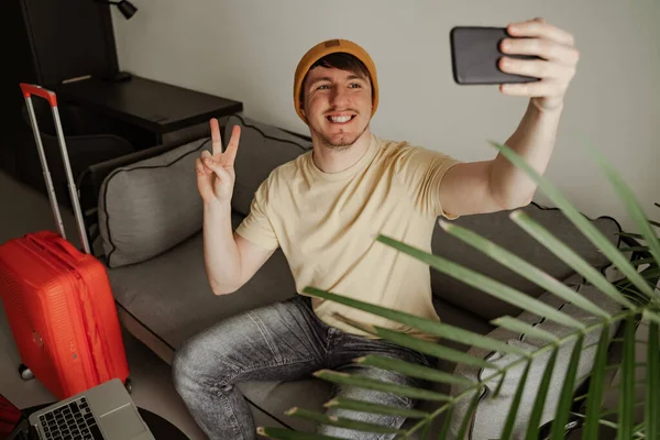 Un viajero joven está sentado en el sofá y haciendo selfie en la habitación de hotel en las vacaciones de verano. Hombre fresco en ropa casual con equipaje — Foto de Stock