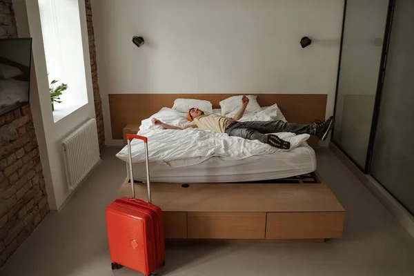 Young man traveler with luggage jumping on bed in hotel room on summer vacation. Cool male in casual clothing