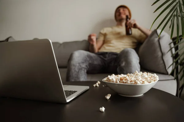 Közelről, fiatal turista sárga kalapban sört iszik popcornnal, filmet néz laptopon a hotelszobában. Férfi vakáción — Stock Fotó