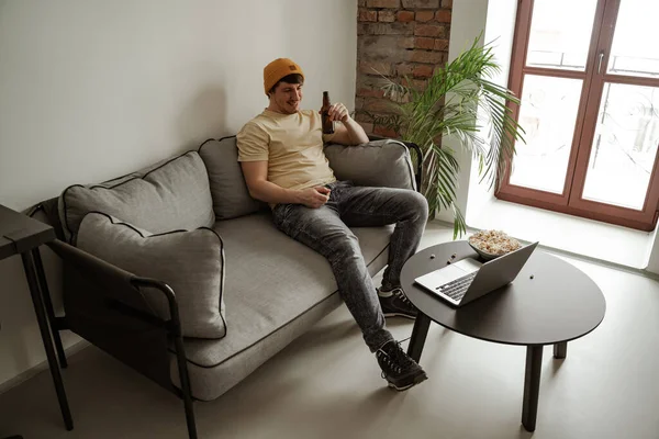 Joven turista en sombrero amarillo bebiendo cerveza con maíz pop, viendo películas en el portátil en la habitación de hotel. Hombre de vacaciones — Foto de Stock