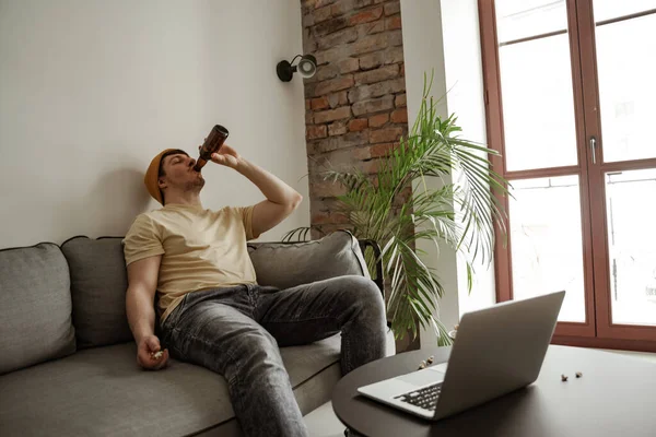 Joven turista en sombrero amarillo bebiendo cerveza con portátil en la habitación de hotel, hombre de vacaciones — Foto de Stock
