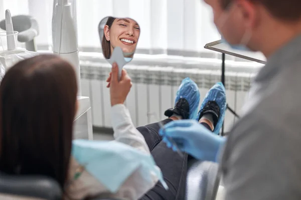 Happy young woman smiling checking out her perfect healthy teeth in the mirror, at the dentist office