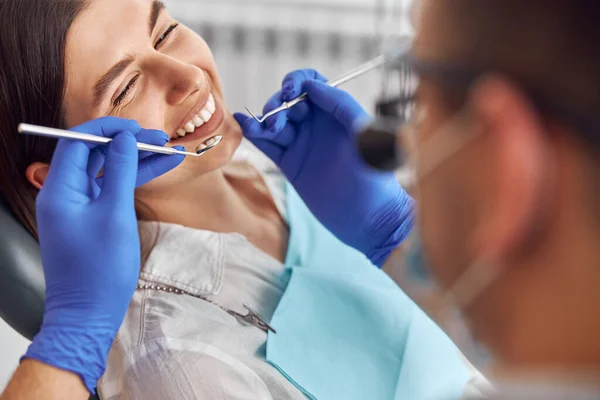 Dentista positivo alegre e cliente em odontologia. Paciente feminina no procedimento odontológico, médica usando instrumentos dentários na clínica odontológica moderna, de perto. — Fotografia de Stock