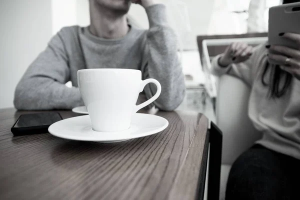 Egy csésze fehér kávé elöl. Homályos pár a háttérben. Férfi és nő egy romantikus randin a kávézóban. Kávéház. Egy nő tartja az okostelefonját és ír valamit, amikor a férfi arra vár, hogy befejezze. — Stock Fotó