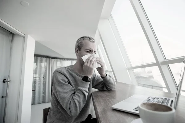 Молодой человек чихает в кофейне. Оставайтесь дома, если вы больны фрилансер работает над своим ноутбуком в кафе или корворкинг дует в нос с помощью бумажной салфетки. Пандемия — стоковое фото