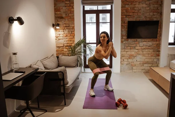 Entrenamiento en casa. Deportes y vida cotidiana. Una joven entrena con una banda elástica. Lleva leggins y usa pesas y una esterilla de yoga. — Foto de Stock