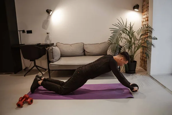 Workout at home. Sports and everyday life. Young man doing exercises on the press with a roller. He is dressed in a tracksuit, there are dumbbells and a yoga mat in the background — Stock Photo, Image