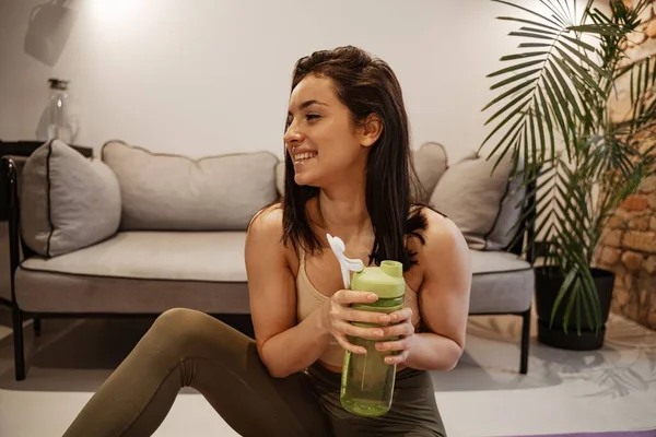 La mujer está haciendo ejercicio en casa. Joven modelo femenina entra para los deportes en el apartamento, ella tiene un agitador de proteínas con leche en las manos. — Foto de Stock