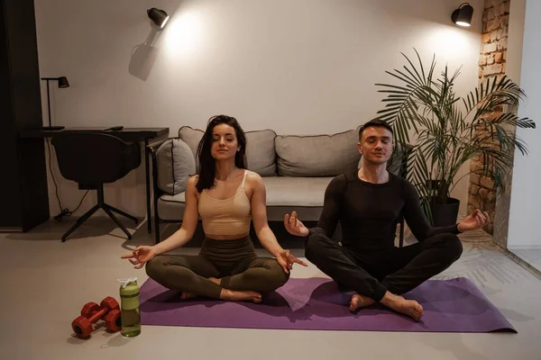 Yoga para dos en casa concepto. Muschina joven y una mujer se sientan en asanas en una esterilla de yoga en un apartamento y meditan. — Foto de Stock