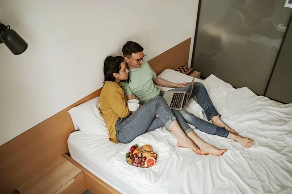 Preciosa pareja viendo películas con sentimientos románticos, sentada en la cama con café y plato de croissant y frutas frescas. Familia feliz mudándose a nuevo apartamento — Foto de Stock