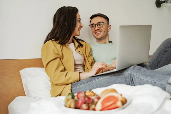 ロマンチックな気持ちで映画を見て素敵なカップル,クロワッサンと新鮮な果物のコーヒーとプレートとベッドの上に座って.幸せな家族は新しいアパートに引っ越します — ストック写真