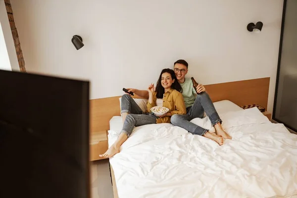 Preciosa pareja con plato de pop conr mientras ve la película en la televisión, sentado en la cama. Familia joven feliz se mudó a nuevo apartamento — Foto de Stock