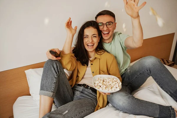 テレビで映画を見ながらポップコーンを投げ素敵なカップルは、ベッドの上に座っている。幸せな若い家族は新しいアパートに移動しました — ストック写真
