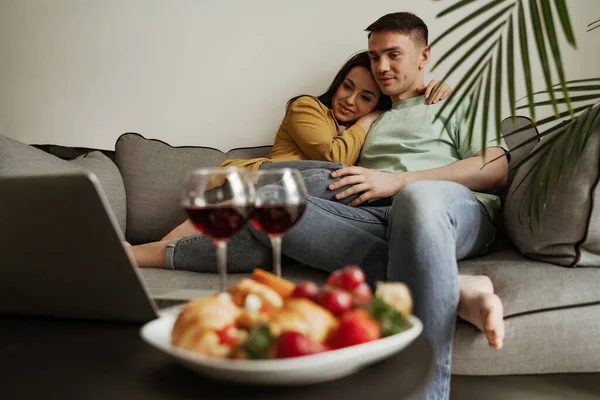 Υπέροχο ζευγάρι βλέποντας ταινία με κρασί και πιάτο κρουασάν και φράουλα, κάθεται στον καναπέ στο σαλόνι. Ευτυχισμένη οικογένεια μετακομίζει σε νέο διαμέρισμα — Φωτογραφία Αρχείου