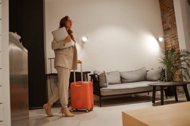 Otelde çalışan genç bir iş kadını. İş gezisi kavramı. Şık bir iş elbisesi ve turuncu bavulu olan kızıl saçlı bir kadın model otele giriş yapıyor..