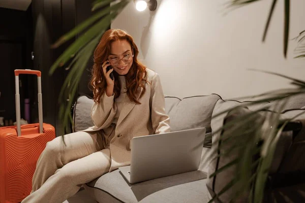 Una joven empresaria trabaja en el hotel. Concepto de viaje de negocios. Modelo femenino de pelo rojo muy ocupado en elegante traje de negocios y maleta naranja trabajando en el ordenador portátil durante su viaje de negocios. — Foto de Stock