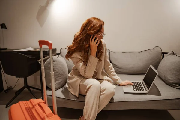 Una joven empresaria trabaja en el hotel. Concepto de viaje de negocios. Modelo femenino de pelo rojo muy ocupado en elegante traje de negocios y maleta naranja trabajando en el ordenador portátil durante su viaje de negocios. — Foto de Stock