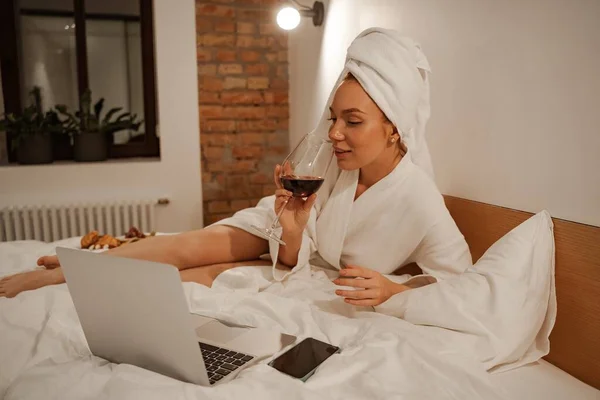 La mujer está vestida con una bata y una toalla blanca en la cabeza. Una joven estudiante pelirroja disfrutando de una copa de vino en casa después de un duro día de trabajo — Foto de Stock
