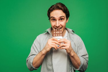 Genç adam yeşil arka planda izole edilmiş çikolata yemekten hoşlanıyor.