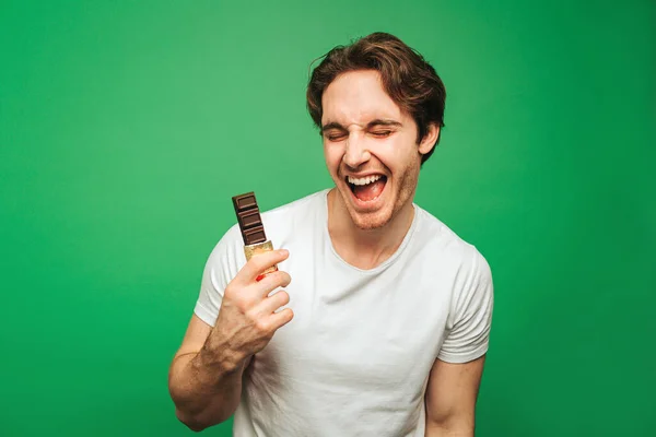 Jongeman houdt chocolade bar en geniet ervan, geïsoleerd op groene achtergrond — Stockfoto