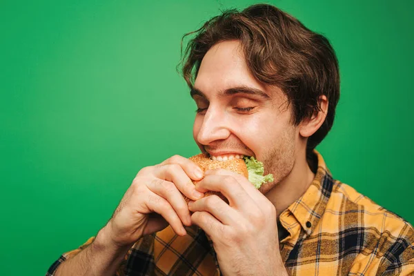 Genç adam yeşil arka planda izole bir şekilde hamburger yiyip eğleniyor. — Stok fotoğraf