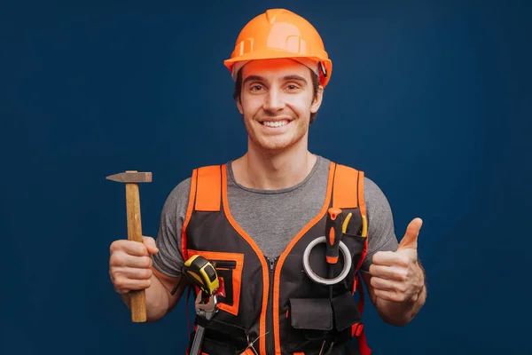 Builder in a helmet with a hammer is smiling and gives thumb up, isolated over blue background — Stock Photo, Image