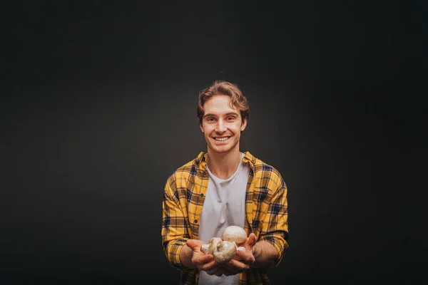 Giovane uomo biondo in camicia gialla tiene funghi in mano con sorriso, isolato su sfondo nero — Foto Stock