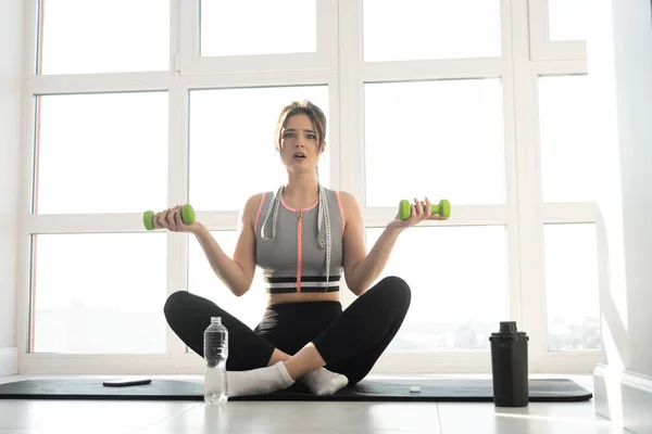 Meglepett nő ül fitness szőnyegen súlyzókkal — Stock Fotó