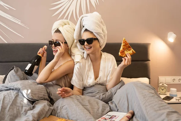 Kadınlar şampanya şişesi ve pizzayı yatakta tutarlar.