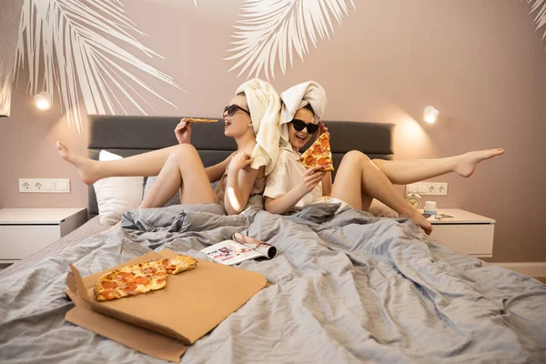 Genç, neşeli kadınlar yatağa uzanıp pizza yiyorlar.