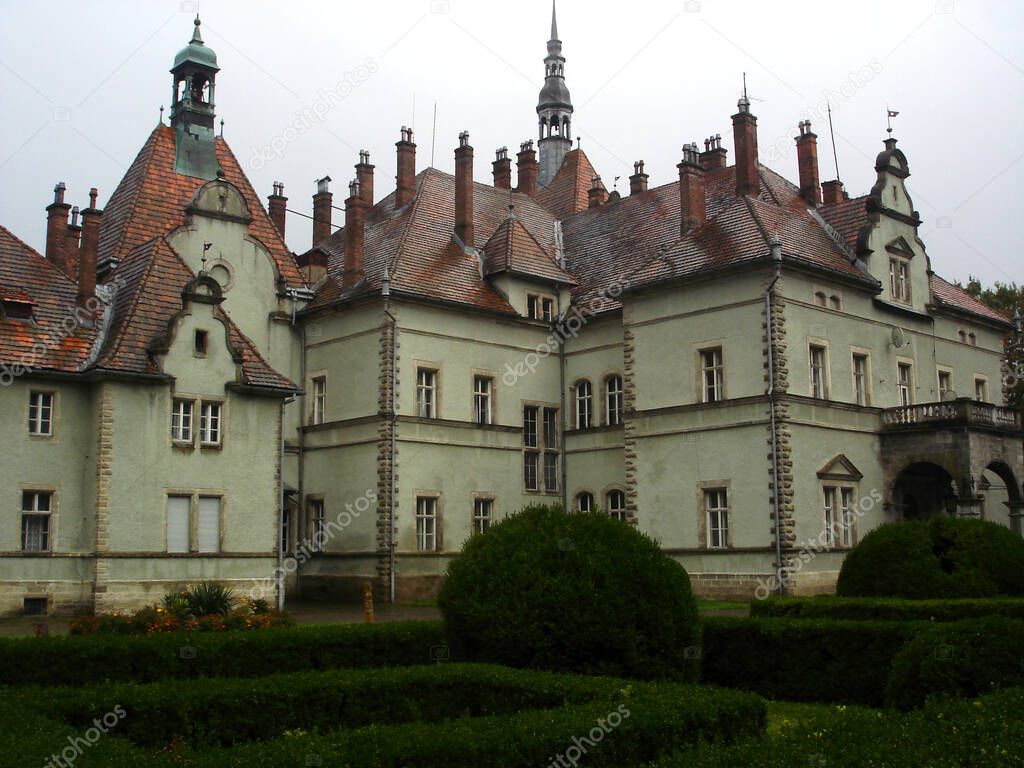 Building is an old castle with a red tiled roof and towers. Bushes and flower beds in the foreground
